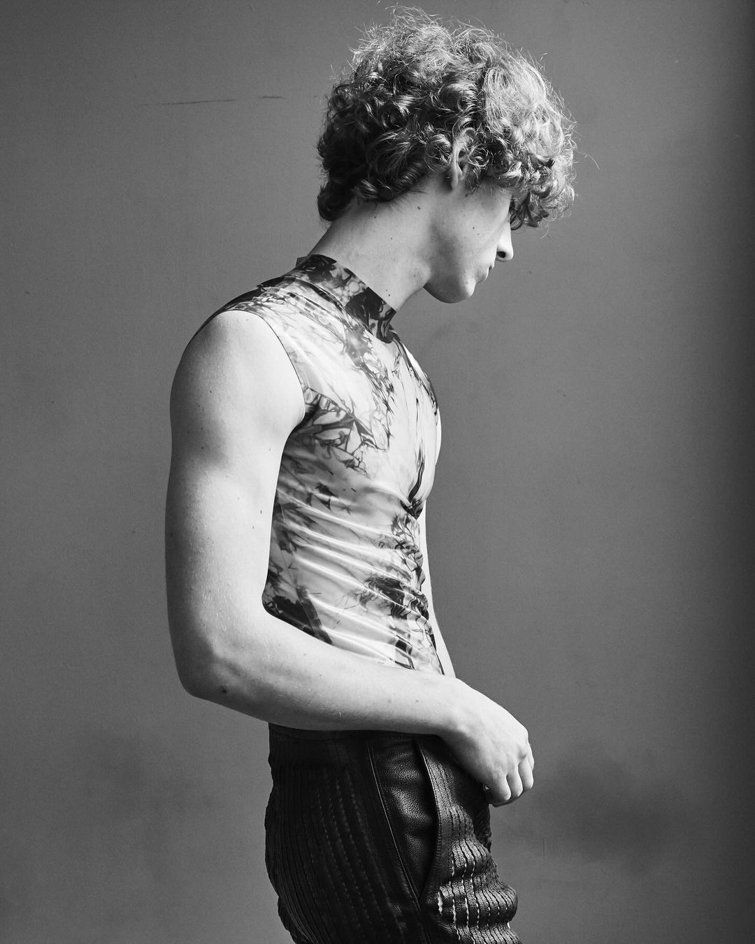 Portrait of an adult male model with curly hair in a patterned top and trousers, looking down against a plain gray backdrop in a studio.