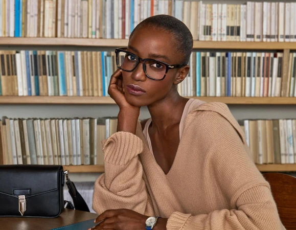 Black woman with short hair wearing thick glasses and a camel v-neck sweater seated in front of a bookshelf.
