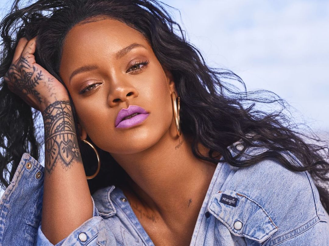 Close-up portrait of Rihanna with striking lilac lipstick, wearing a denim jacket and gold hoop earrings against a blurred sky background.