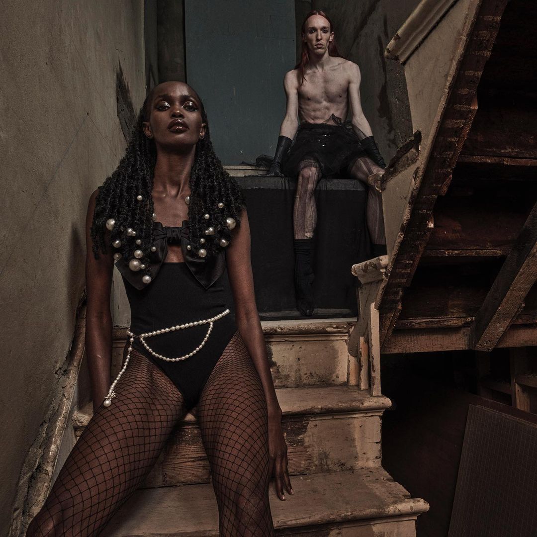 Two fashion models in a grungy indoor setting. A Black woman in a pearl-accented bodysuit and a Caucasian man in a skirt pose on stairs.