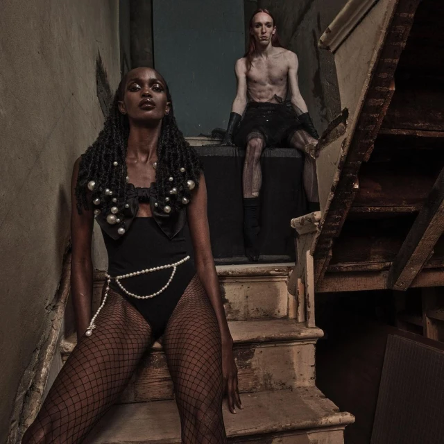 Two fashion models in a grungy indoor setting. A Black woman in a pearl-accented bodysuit and a Caucasian man in a skirt pose on stairs.