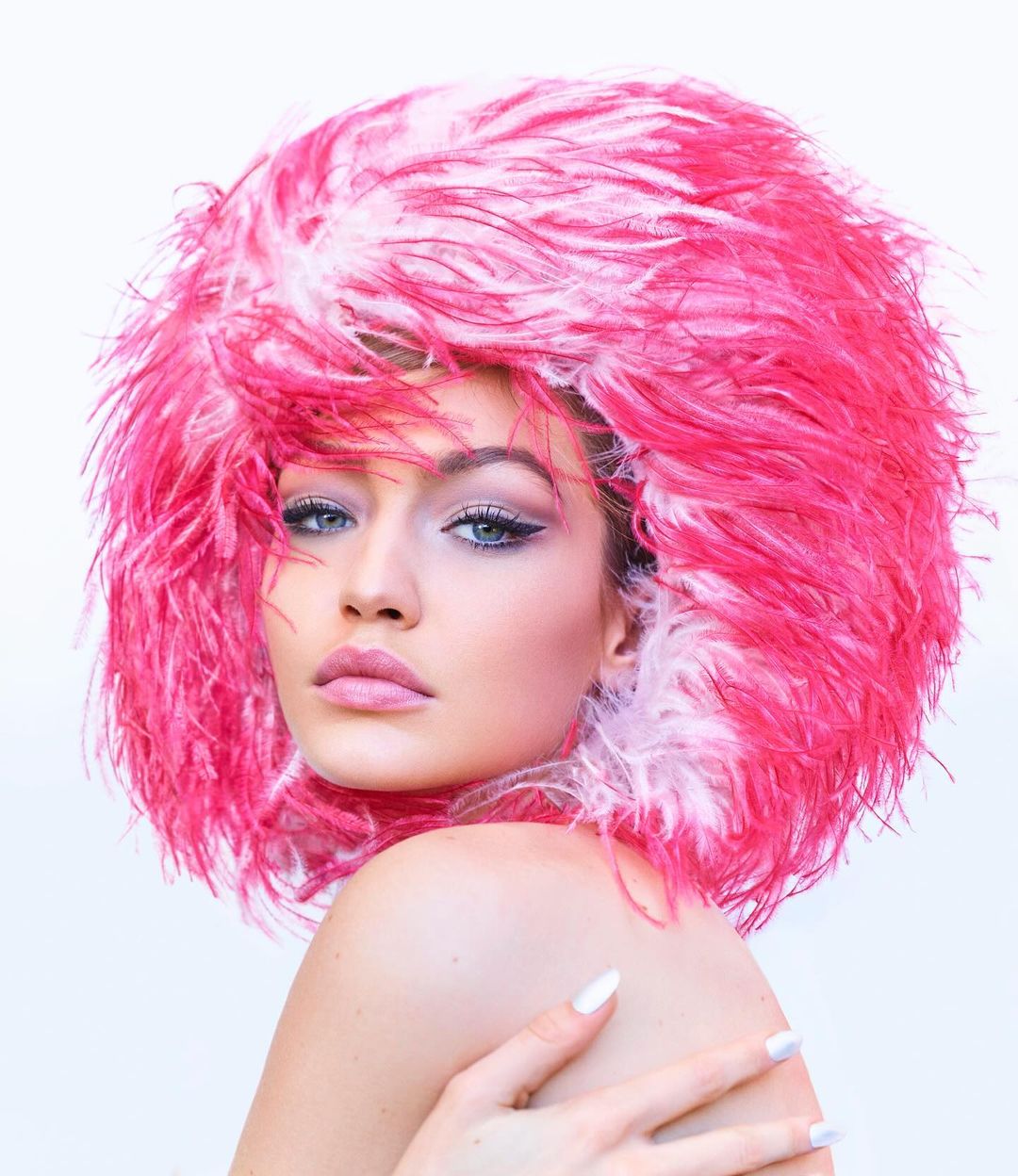 Close-up portrait of a woman with blue eyes and pink lips wearing a large, bright pink feathered hat against a white backdrop.