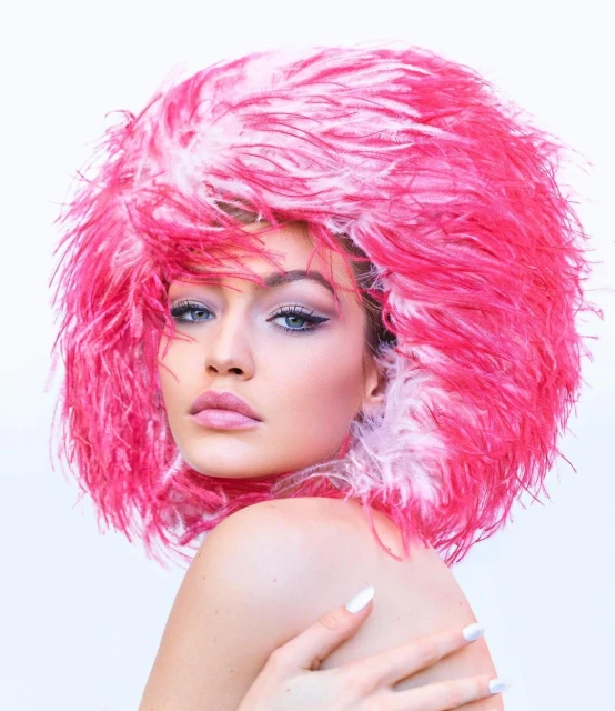 Close-up portrait of a woman with blue eyes and pink lips wearing a large, bright pink feathered hat against a white backdrop.