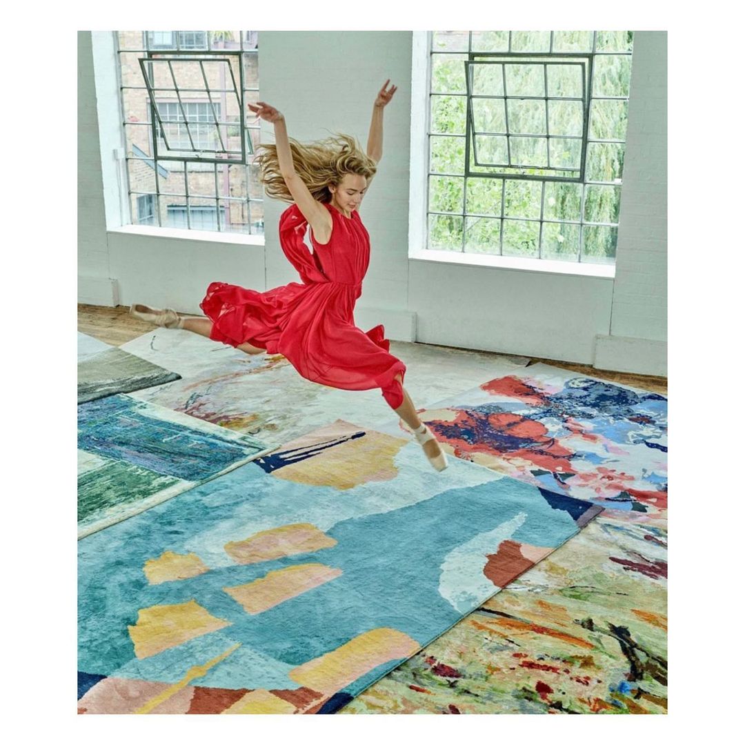 Woman in red dress leaps mid-air above colorful area rugs in a light-filled room with large windows, showcasing a dynamic and artistic scene.