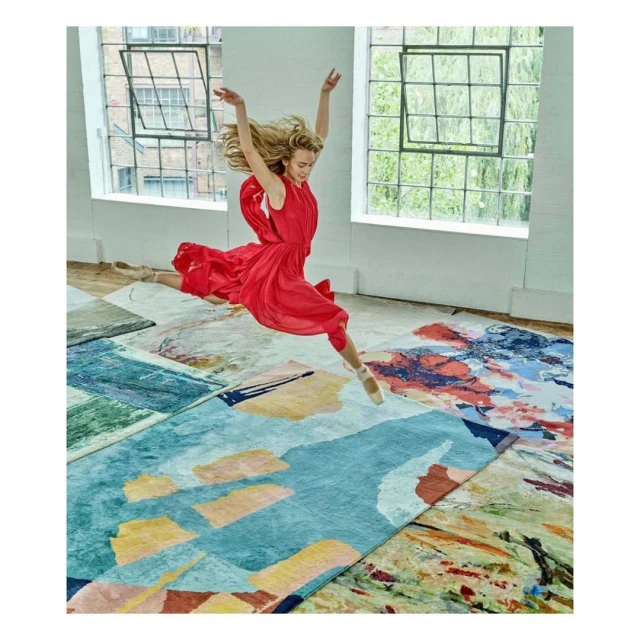Woman in red dress leaps mid-air above colorful area rugs in a light-filled room with large windows, showcasing a dynamic and artistic scene.