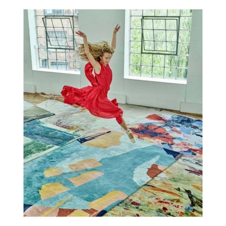 Woman in red dress leaps mid-air above colorful area rugs in a light-filled room with large windows, showcasing a dynamic and artistic scene.