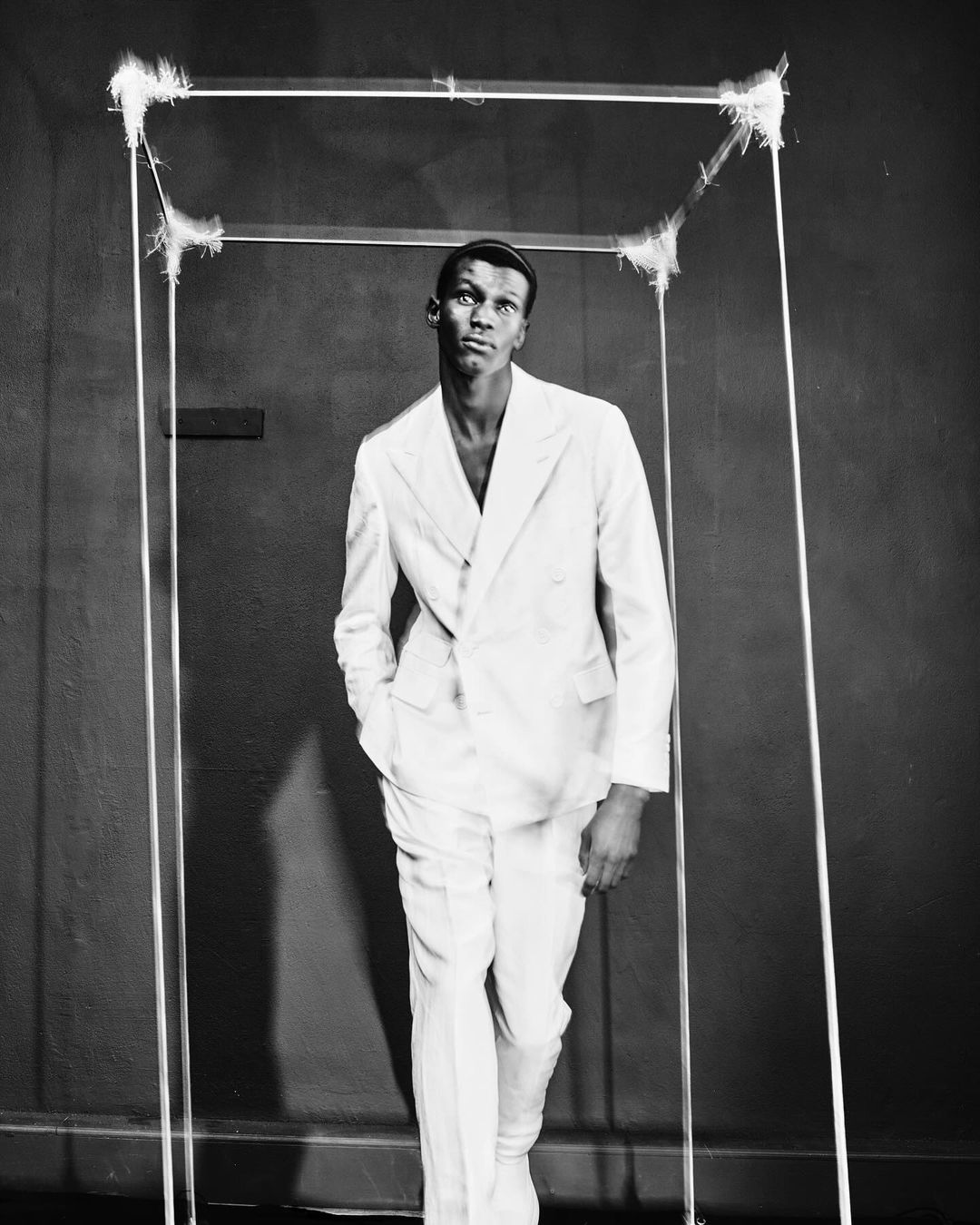 Monochrome portrait of an African-American man in a white suit, standing inside a deconstructed square frame art piece against a dark background.