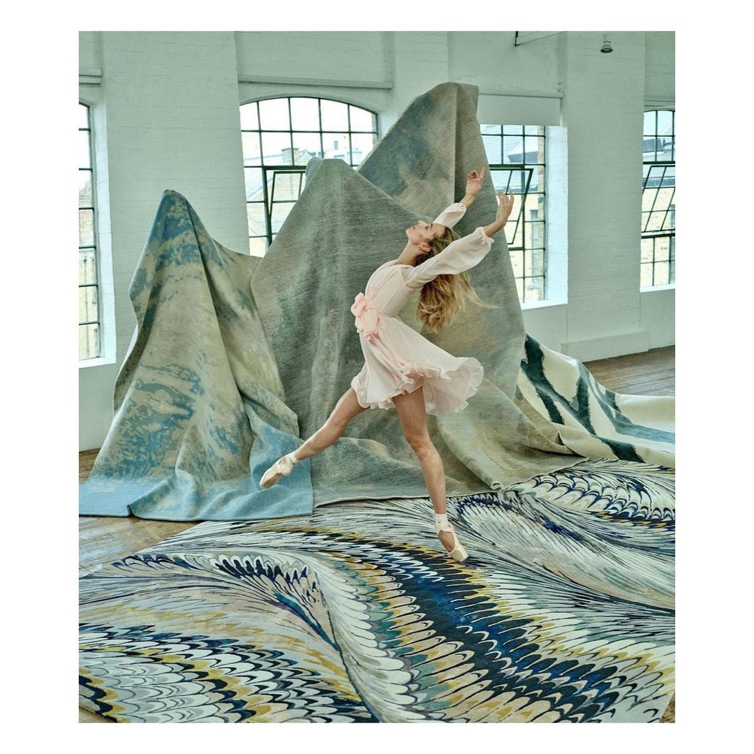 A ballerina leaps across a marble-patterned rug, in front of folded textile mountains, inside a brightly lit industrial space.