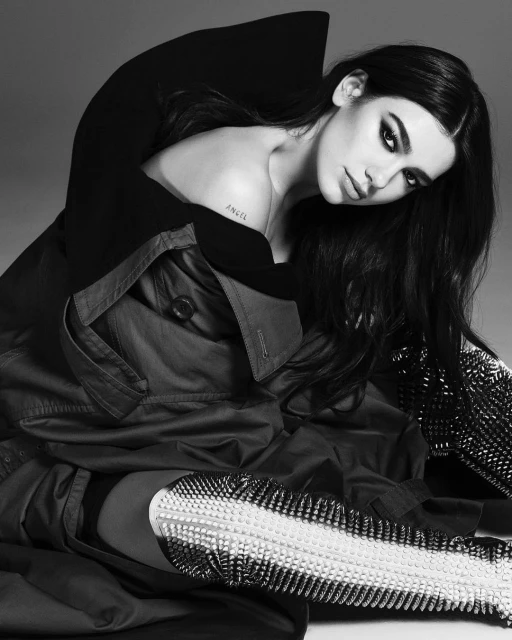 Black and white portrait of a woman with dark hair wearing shiny, beaded boots and a dark coat in a studio setting.