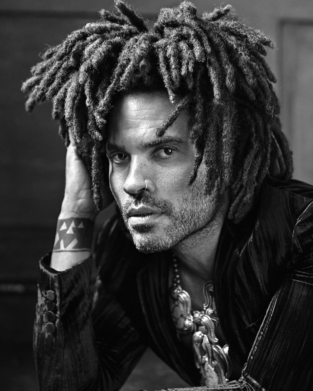 Black and white portrait of a stylish man with dreadlocks wearing a velvet jacket and ornate pendant necklace looking at the camera.