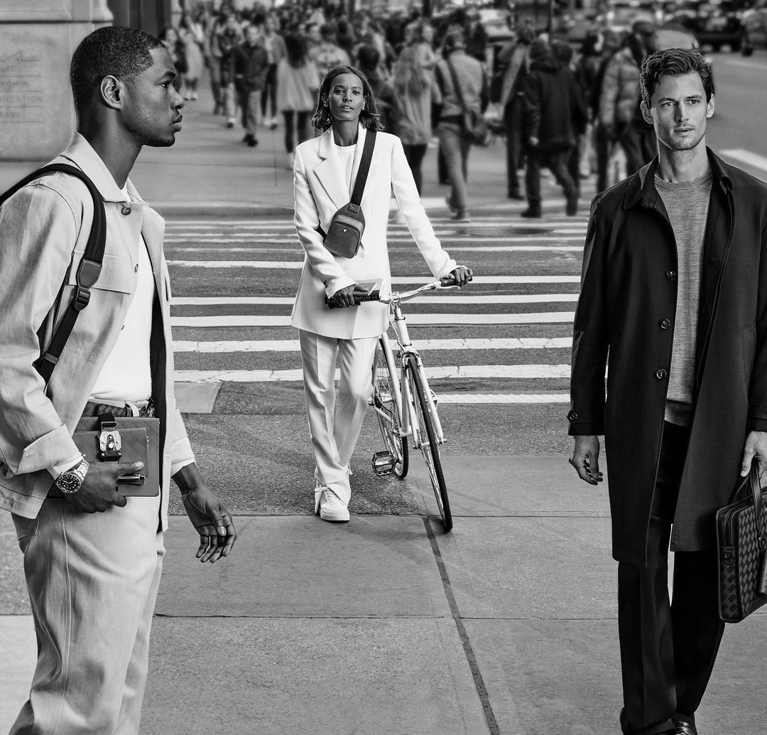 Three people in black and white in the city. A woman is walking her bike on the crosswalk in front of other people.