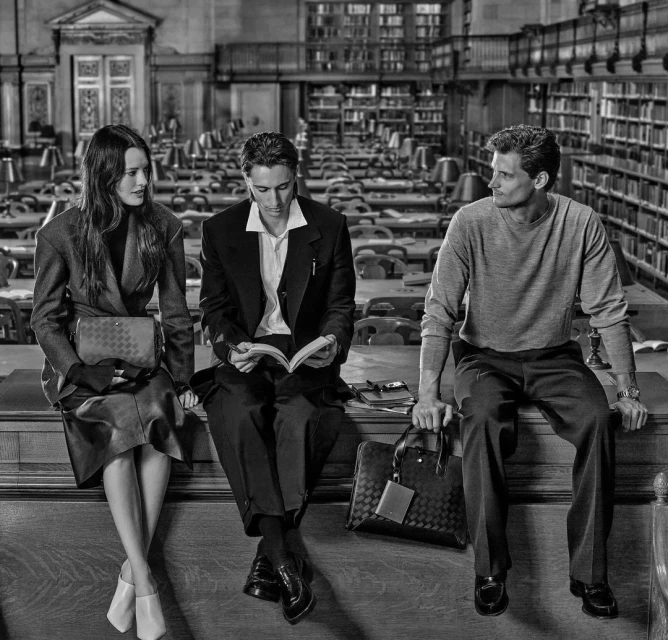 Three people sit on a ledge in a library setting. A woman wears a coat, a man in a suit reads, and another man in a sweater looks to the side.