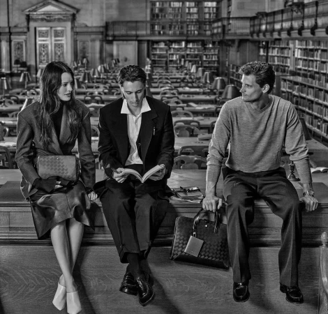 Three people sit on a ledge in a library setting. A woman wears a coat, a man in a suit reads, and another man in a sweater looks to the side.