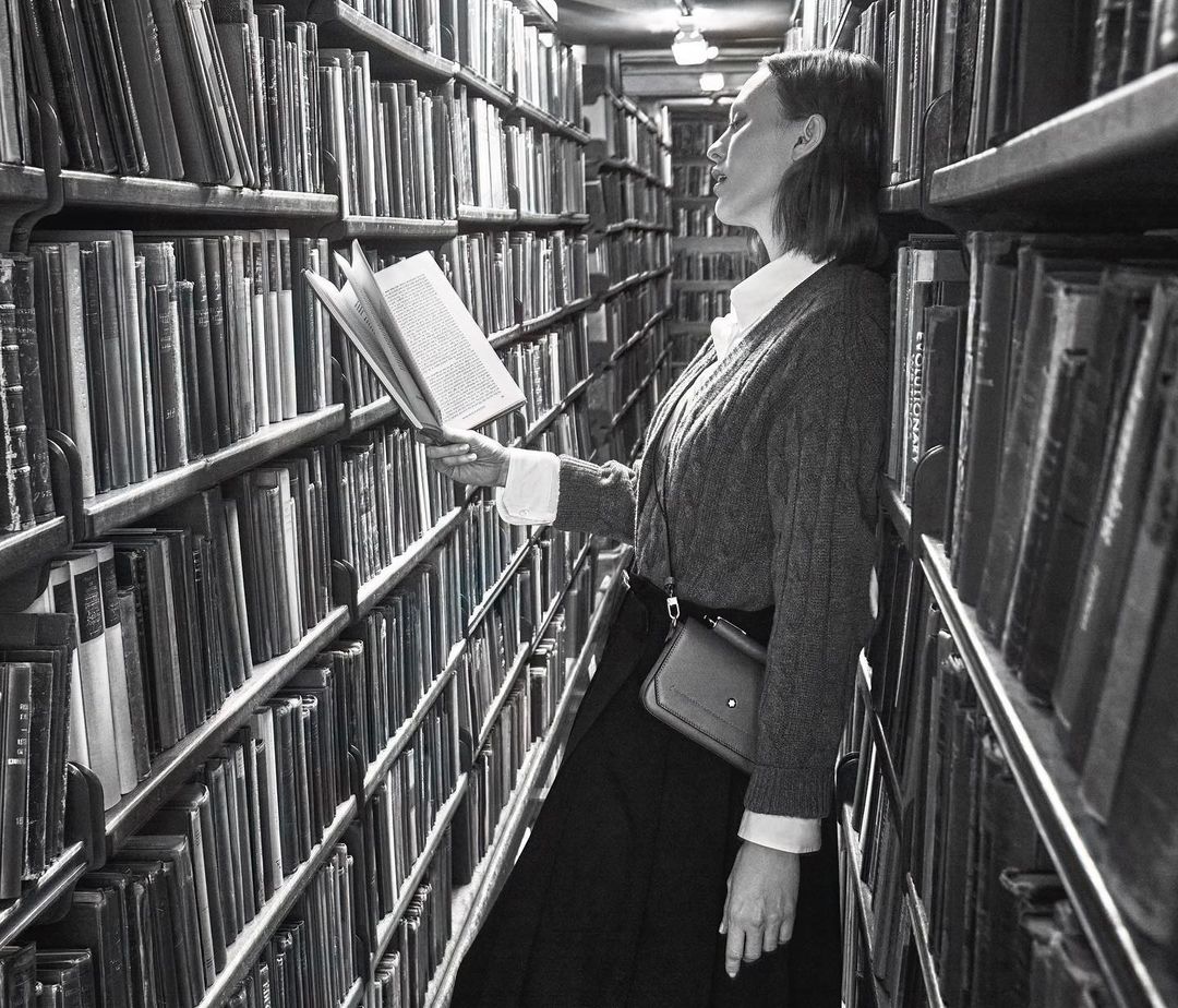 Woman in library reading with shelves of books behind her in a monochromatic portrait-style photograph, offering a vintage, nostalgic feel.