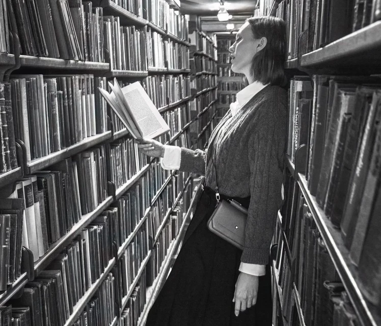 Woman in library reading with shelves of books behind her in a monochromatic portrait-style photograph, offering a vintage, nostalgic feel.