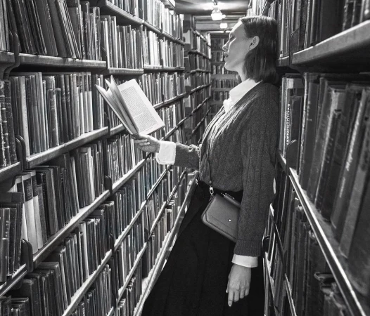 Woman in library reading with shelves of books behind her in a monochromatic portrait-style photograph, offering a vintage, nostalgic feel.