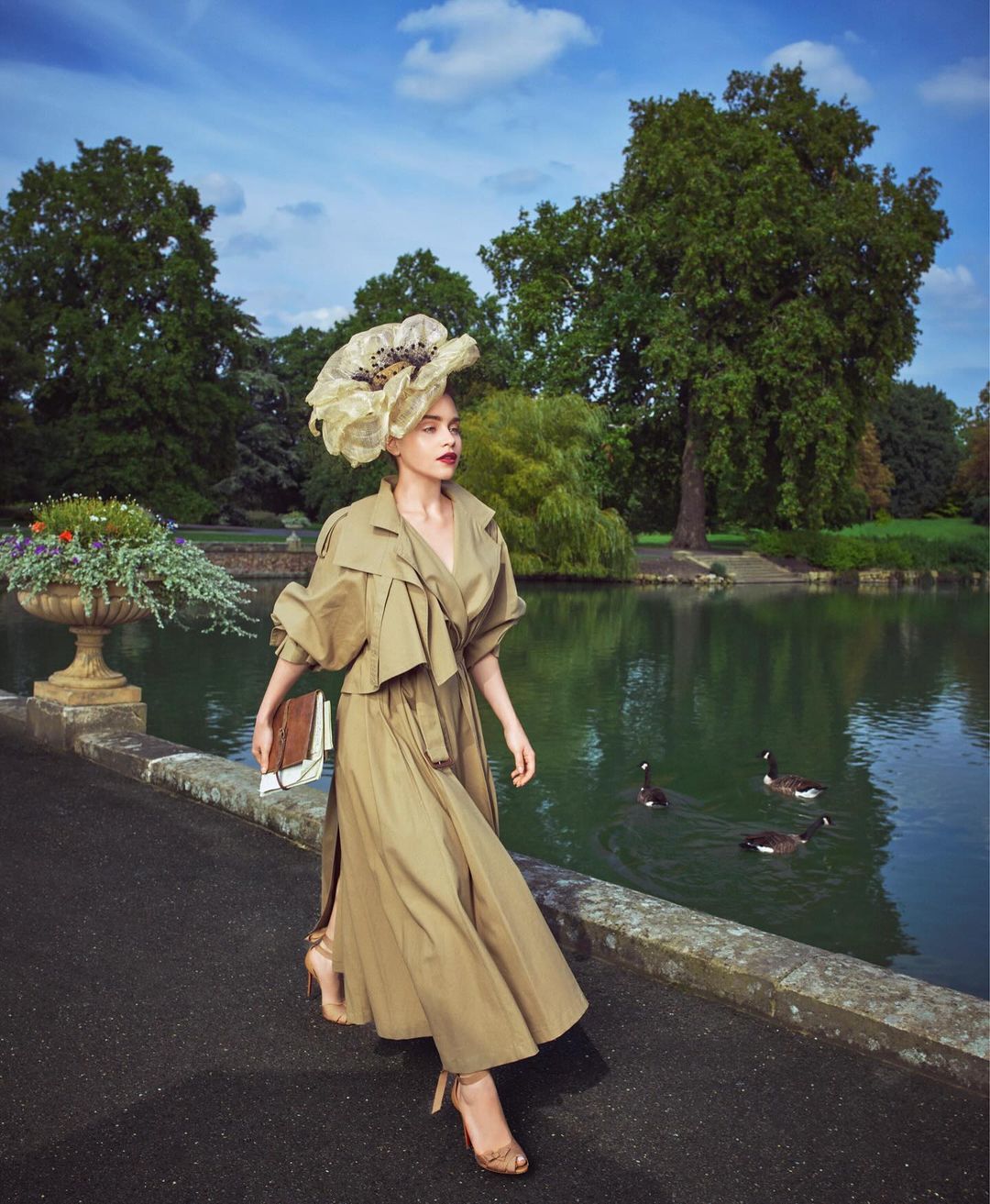 Woman in a khaki coat and large decorative hat walking on a bridge over a pond with ducks and trees in the background on a sunny day.