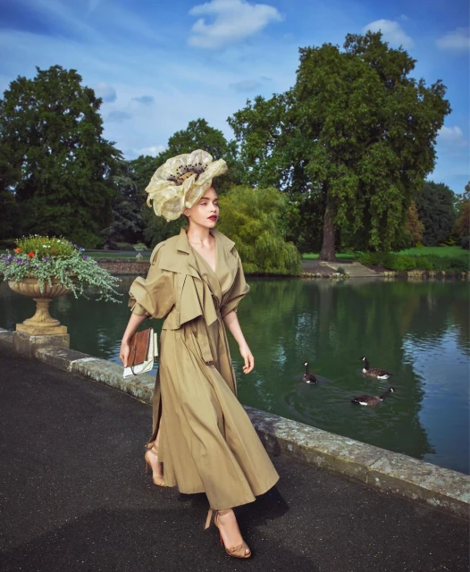 Woman in a khaki coat and large decorative hat walking on a bridge over a pond with ducks and trees in the background on a sunny day.