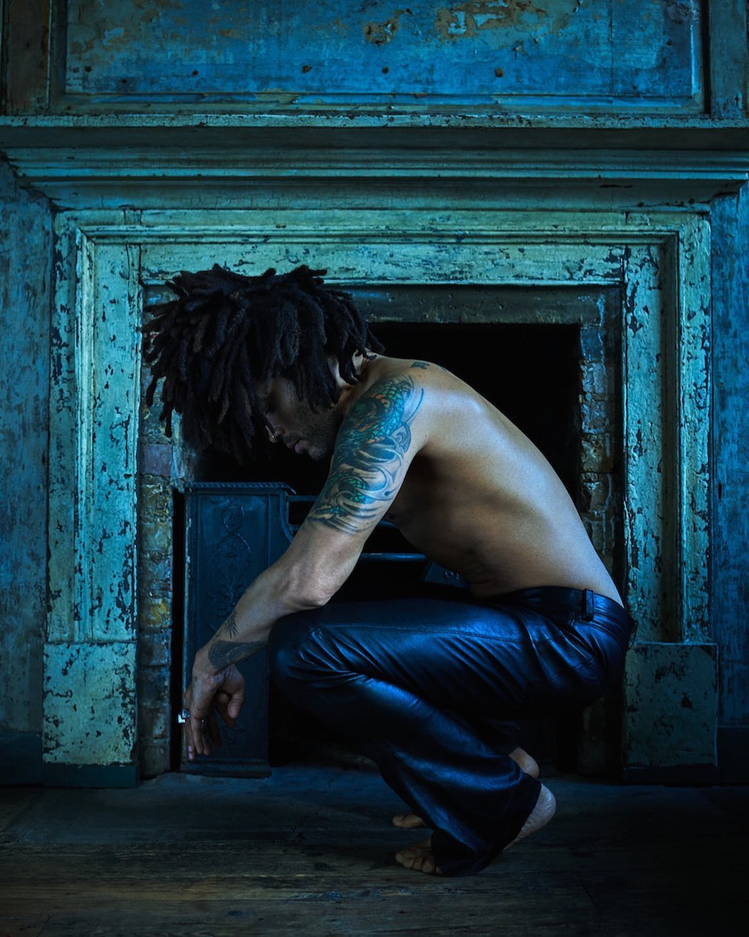 A shirtless man with dreadlocks and tattoos crouches in a dark interior with a distressed fireplace and a blue color cast, creating a moody image.