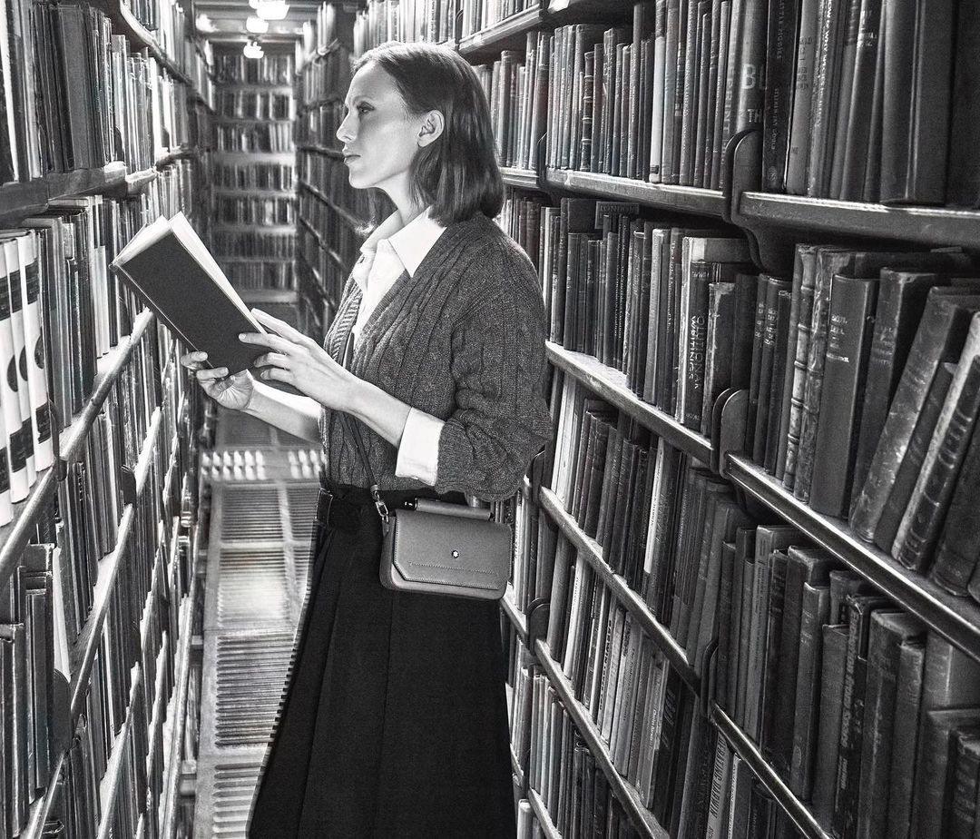 A woman stands in a black and white library setting, holding and reading an open book, surrounded by shelves filled with books.
