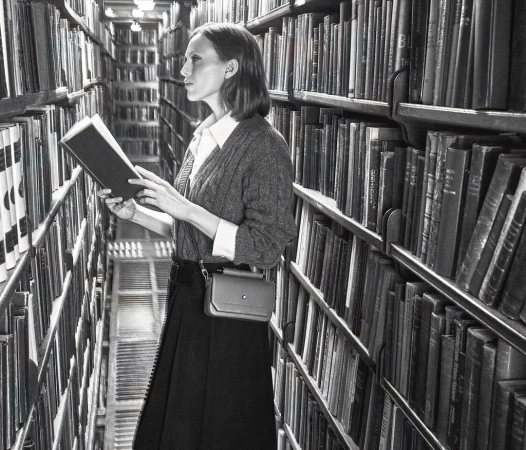 A woman stands in a black and white library setting, holding and reading an open book, surrounded by shelves filled with books.