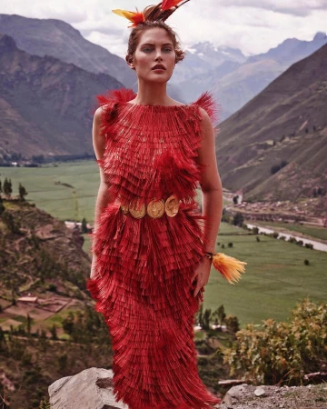 Woman in a layered red dress with gold accents and feathers poses in front of a mountain range landscape with a green valley.