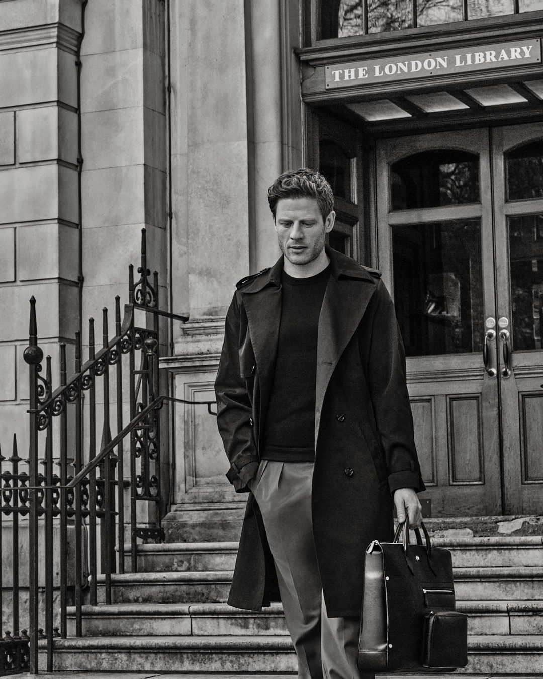 A stylish adult male in a dark trench coat and slacks walks down stone steps while holding a briefcase in front of The London Library.