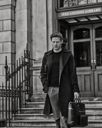 A stylish adult male in a dark trench coat and slacks walks down stone steps while holding a briefcase in front of The London Library.