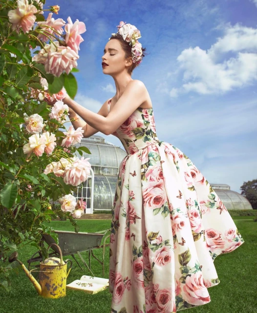 Woman in floral dress stands in garden with roses and greenhouse. Wearing floral headpiece, she picks roses near vintage wheelbarrow.