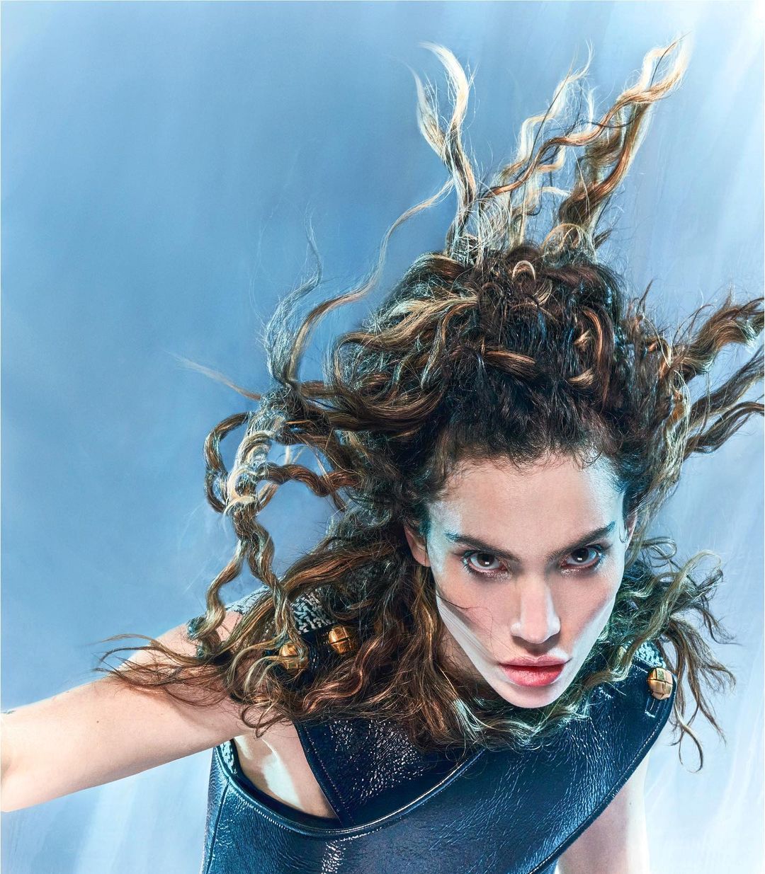 Eye-level shot of a striking brunette woman with curly hair in a dark, glossy top against a soft blue background in a beauty editorial.