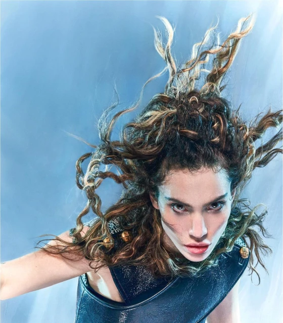 Eye-level shot of a striking brunette woman with curly hair in a dark, glossy top against a soft blue background in a beauty editorial.