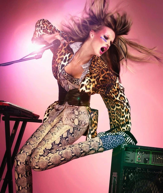 A blonde female musician energetically sings into a microphone while standing near equipment in a photo studio with a pink backdrop.