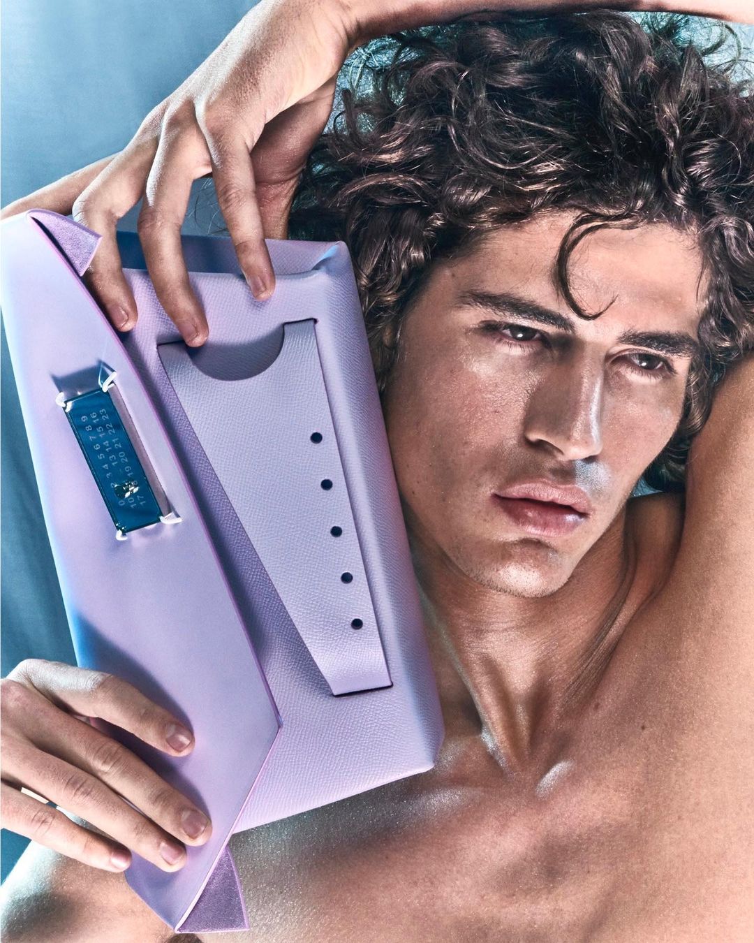 Adult male model poses with a lavender leather clutch bag. Fashion photography showcases men's style and accessories in a studio setting.