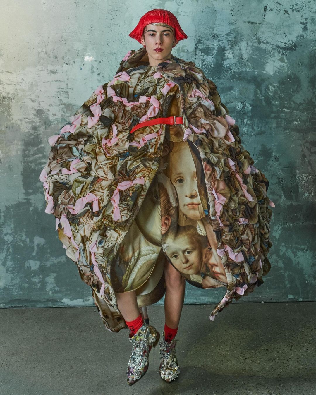 A person in a voluminous, patterned coat with a red belt, standing in front of a textured gray wall, wearing red wig and patterned boots.