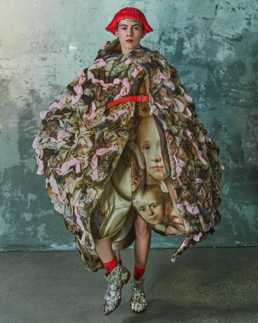 A person in a voluminous, patterned coat with a red belt, standing in front of a textured gray wall, wearing red wig and patterned boots.