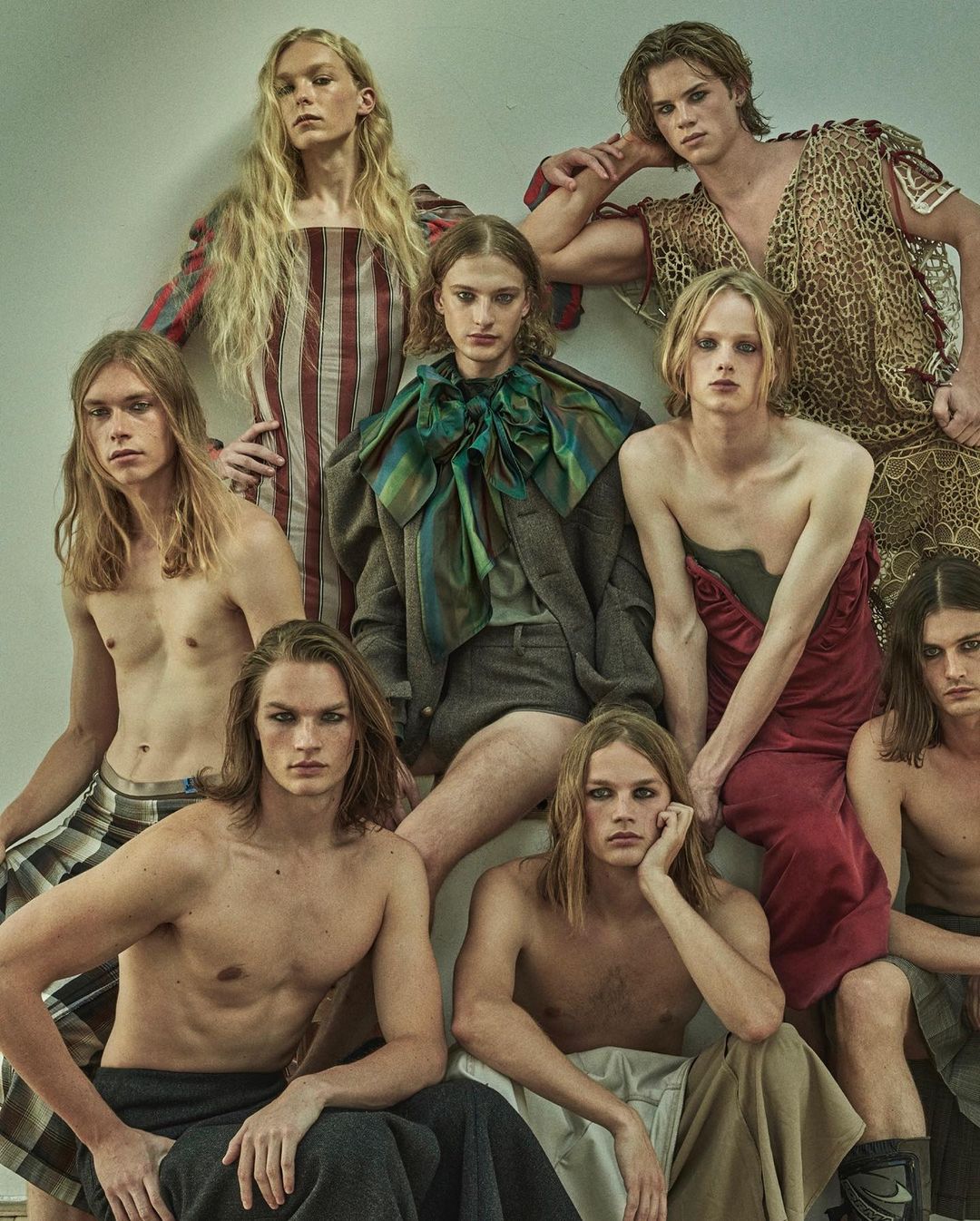 A diverse group of androgynous-looking male fashion models with long hair in various poses and attire in a studio setting.
