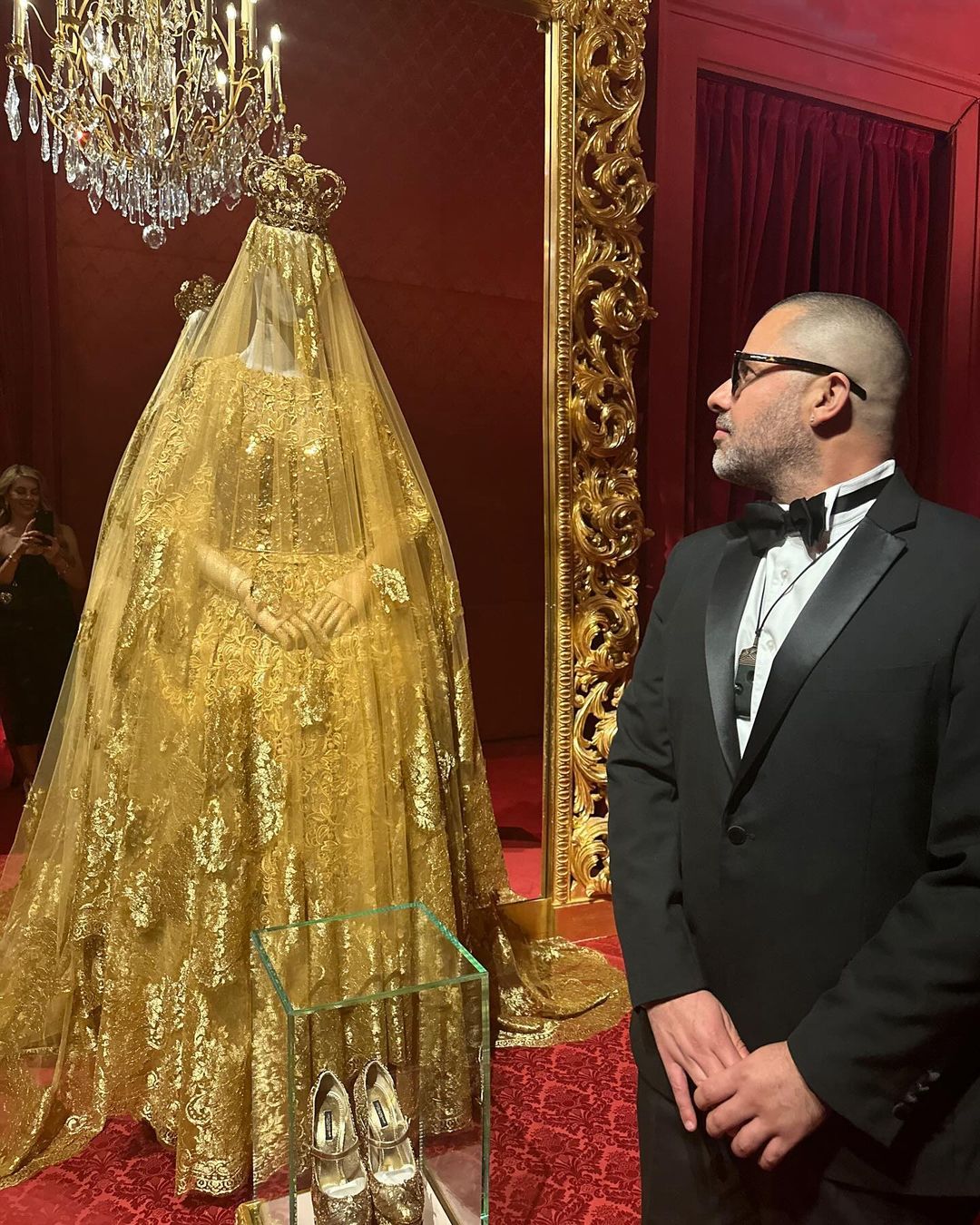 An ornate gold dress with a crown on a mannequin behind glass, with a man in a tuxedo viewing it, in an opulent room.