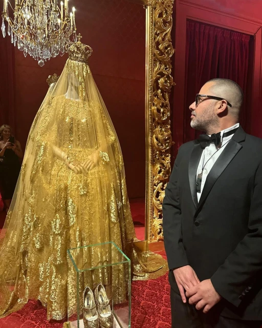 An ornate gold dress with a crown on a mannequin behind glass, with a man in a tuxedo viewing it, in an opulent room.