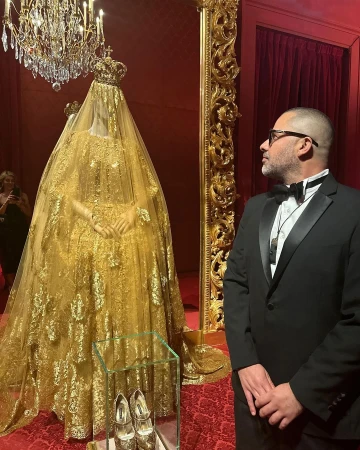 An ornate gold dress with a crown on a mannequin behind glass, with a man in a tuxedo viewing it, in an opulent room.