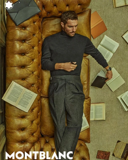 Adult male model lays on a tan leather couch, surrounded by books, wearing a dark gray sweater and gray wool pants, holding a pen.