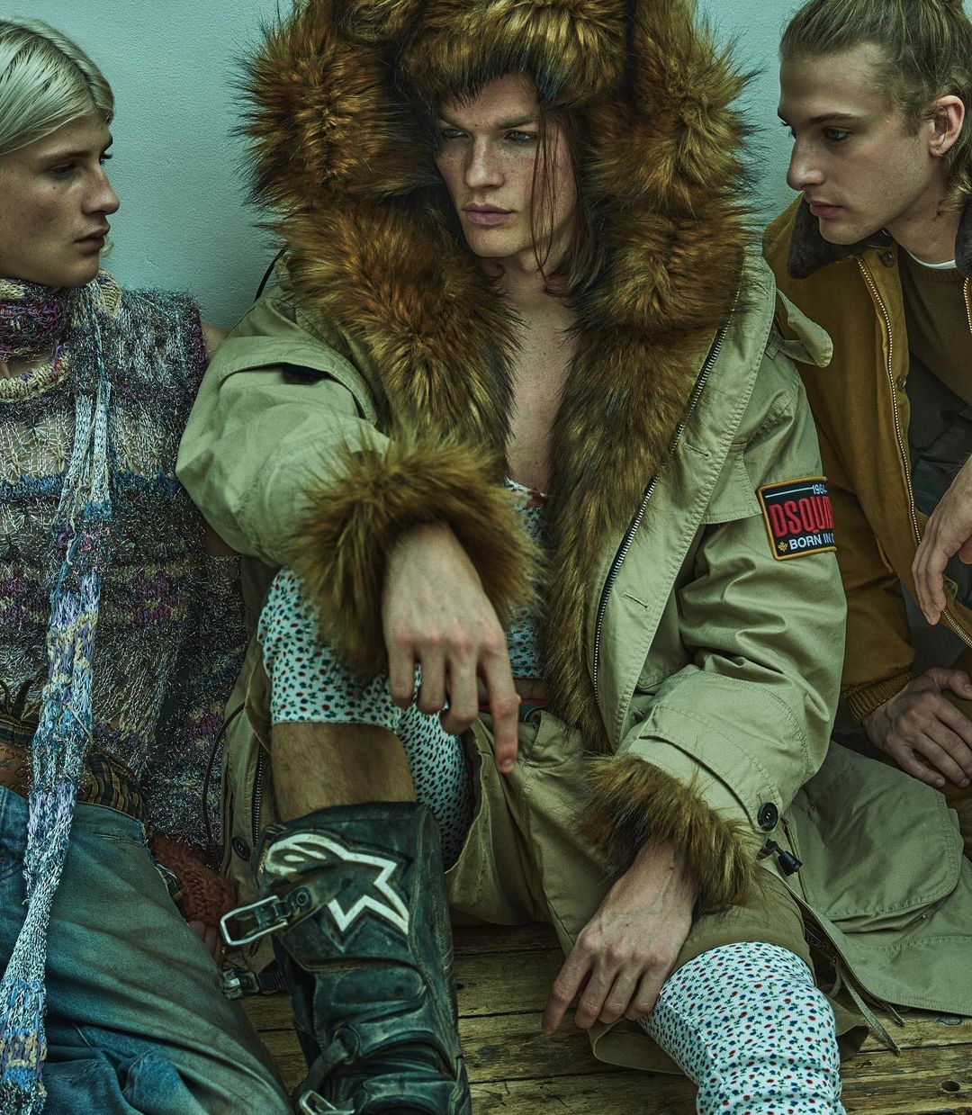 Three models pose indoors, the central figure wears a fur-trimmed hooded coat and high boots in a fashion editorial setting. 