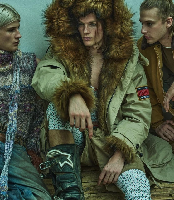 Three models pose indoors, the central figure wears a fur-trimmed hooded coat and high boots in a fashion editorial setting. 