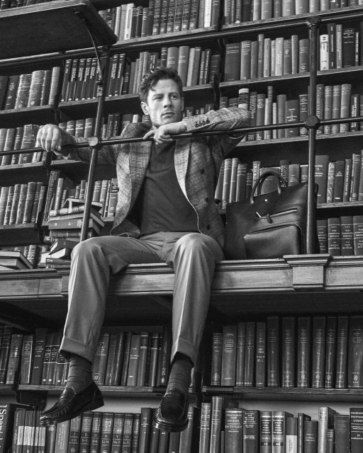 Monochrome portrait of a man sitting on a bookshelf, wearing a jacket and slacks, with a leather briefcase nearby.