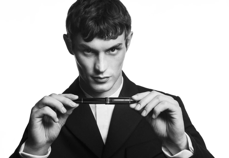 Monochrome portrait of a young adult male holding a black Montblanc pen in front of his chest while wearing a dark suit.