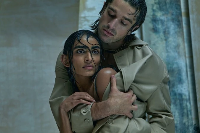 An adult couple in a close indoor portrait, with a fair-skinned man wearing a trench coat embracing a woman in neutral lighting.
