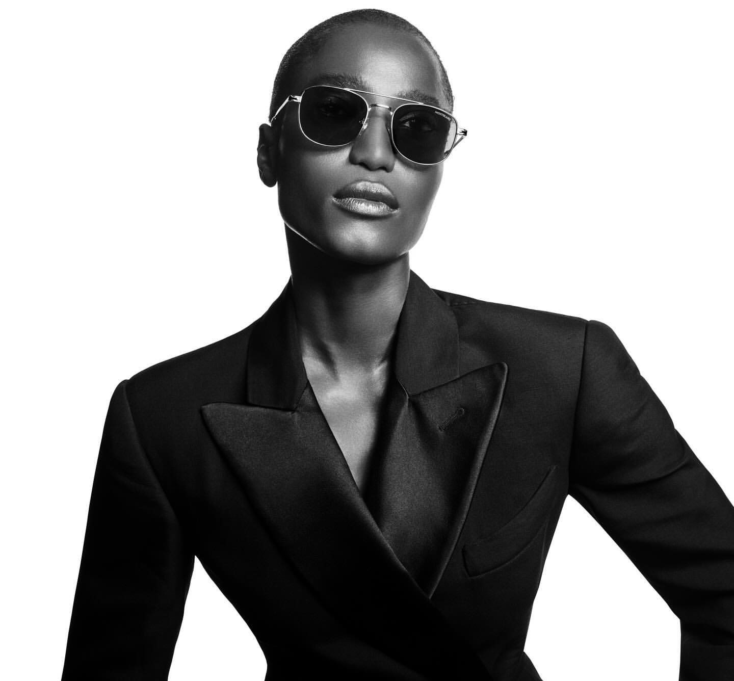 Close-up portrait of a woman wearing sunglasses and a black blazer against a white background in a black-and-white photograph.
