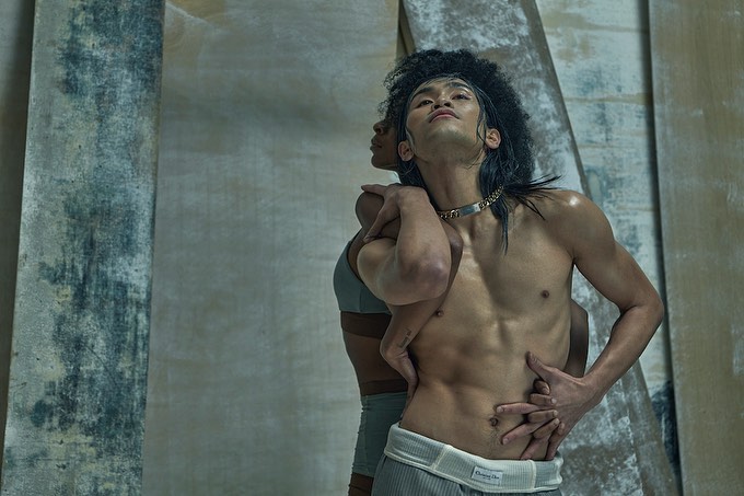 An artistic shot features an Asian male model embraced by another person, showcasing toned physique and unique styling against a textured backdrop.