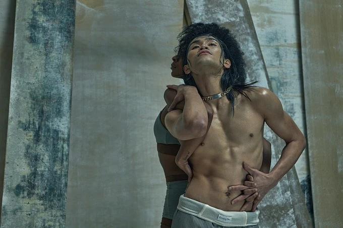 An artistic shot features an Asian male model embraced by another person, showcasing toned physique and unique styling against a textured backdrop.