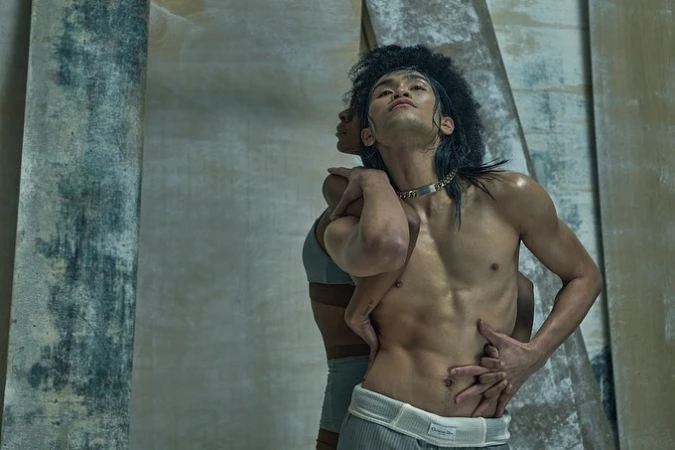 An artistic shot features an Asian male model embraced by another person, showcasing toned physique and unique styling against a textured backdrop.