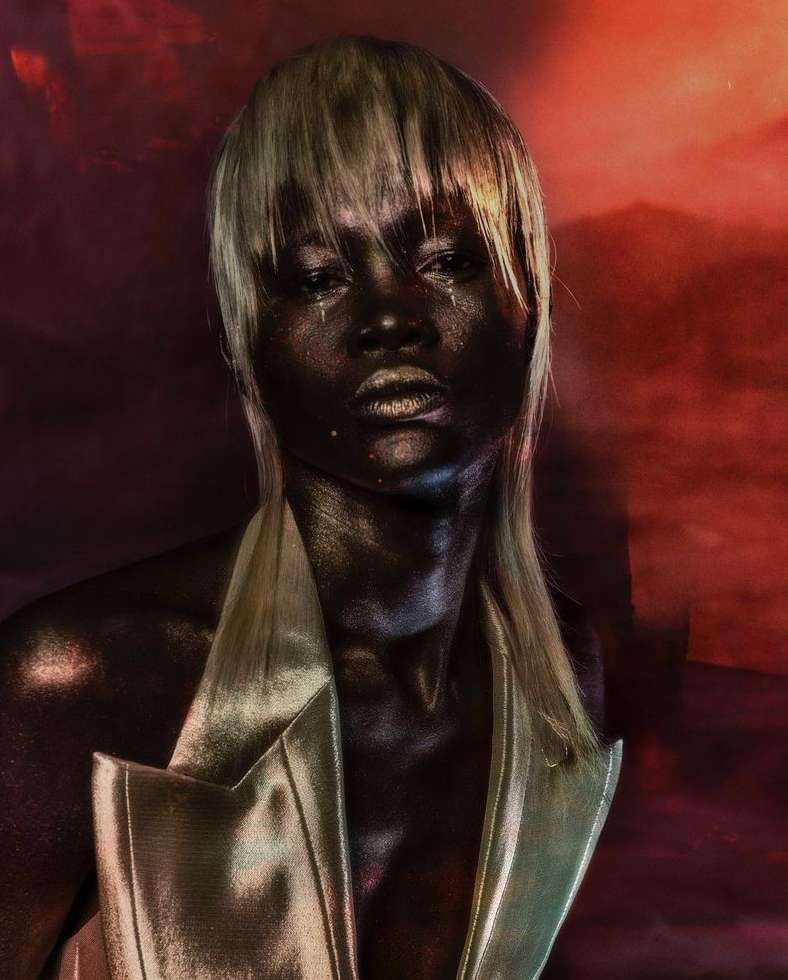 Striking beauty portrait of a dark-skinned woman with platinum blonde hair, metallic gold makeup, and a gold jacket, set against a red background.
